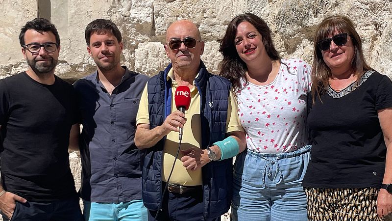 Cinco personas sonríen al aire libre frente a una pared de piedra. Paco Nadal, periodista, sostiene un micrófono de RNE.