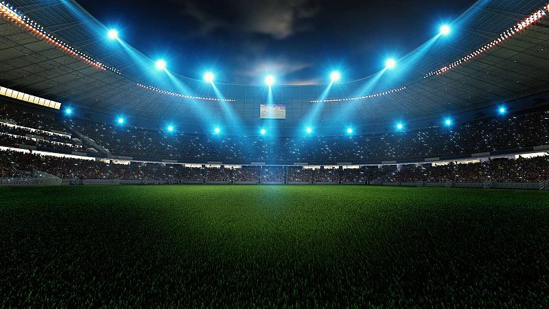 Estadio de fútbol moderno de noche, con campo de césped verde iluminado y gradas llenas de público. Focos azules y pantalla gigante en la distancia.
