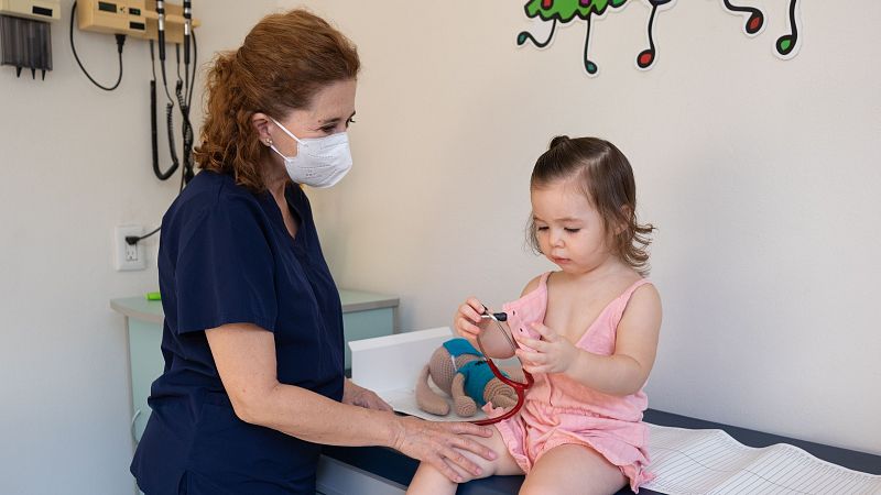 Profesional de la salud interactúa con niña que juega con estetoscopio de juguete en consultorio pediátrico. Ambiente infantil con notas musicales decorativas.