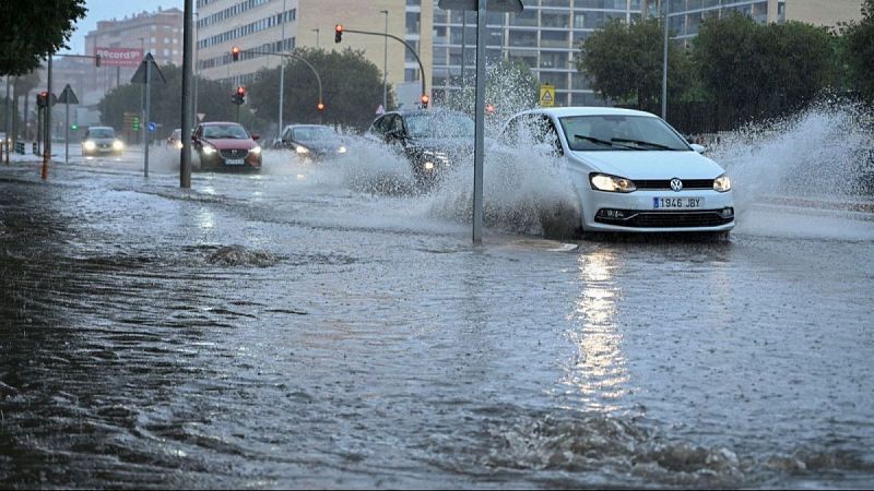 Un Volkswagen Polo blanco, matrícula "1946 JBY", avanza por una calle urbana inundada por fuertes lluvias, con los faros encendidos y generando salpicaduras. Otros coches circulan en la vía anegada bajo un cielo nublado.