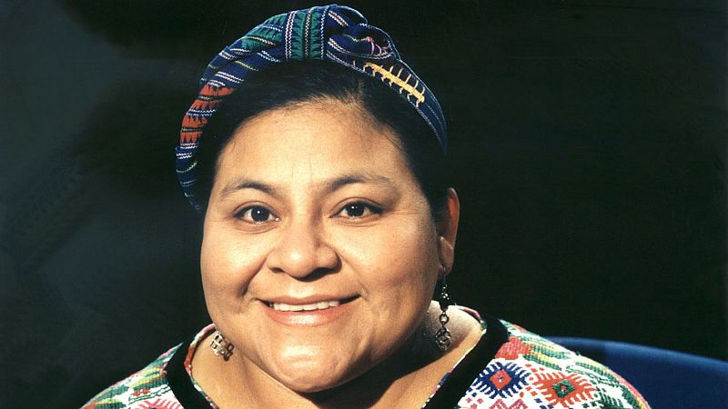 Retrato de Rigoberta Menchú, activista guatemalteca y Premio Nobel de la Paz, luciendo un tocado y huipil tradicionales con bordados geométricos y pendientes de metal oscuro.