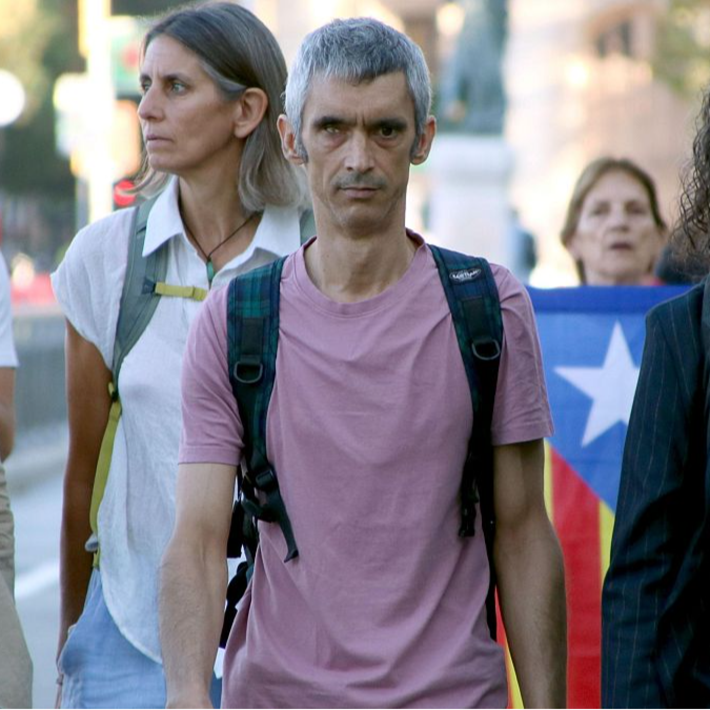 Primer plano de Roger Español con camiseta rosa y mochila de cuadros, acompañado de una mujer y una bandera Estelada al fondo. Expresión seria y ojeras marcadas.