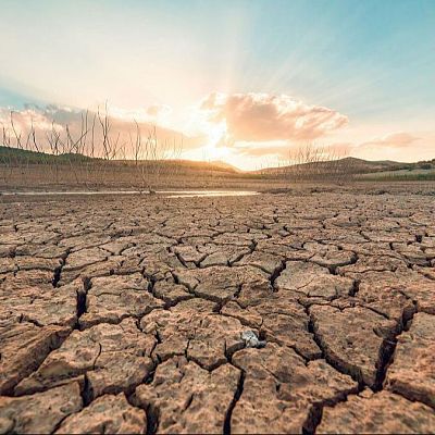 Paisaje africano árido con suelo agrietado, árboles sin hojas y un charco bajo un cielo crepuscular anaranjado y azul, indicando sequía extrema.