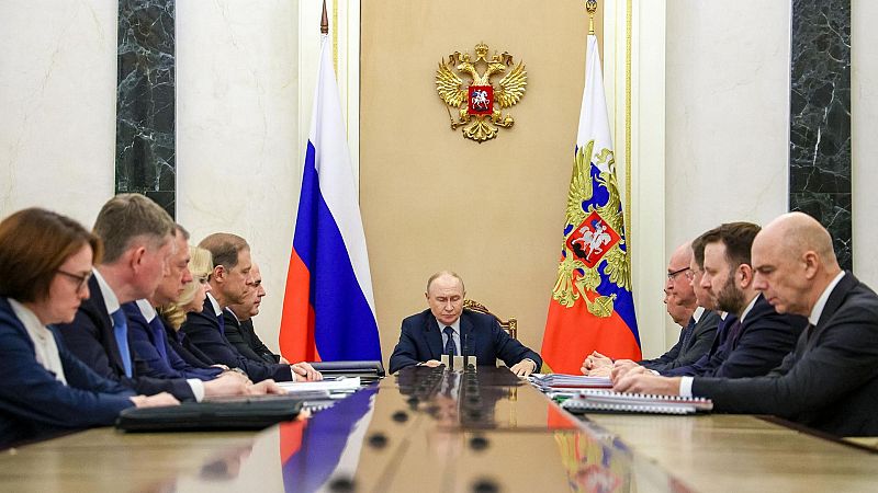 Vladímir Putin, presidente de Rusia, preside una reunión oficial. El águila bicéfala y la bandera rusa adornan la pared detrás de él. Varios asistentes con trajes oscuros se sientan a su alrededor.