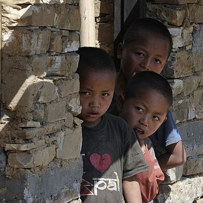 Tres niños asiáticos asoman desde una pared de piedra. El de la izquierda lleva camiseta gris con \"Nepal\" y corazón rojo. Luz solar intensa.