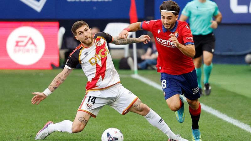 Isi Palazón (Rayo Vallecano #14) y Jon Moncayola (Osasuna #8) compiten por el balón en un partido de fútbol. Se ven detalles de sus camisetas, patrocinadores y un árbitro al fondo.