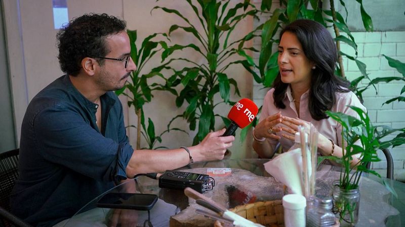 Claudia Ortiz, opositora salvadoreña, es entrevistada por un periodista de Radio Nacional de España en un estudio con plantas. Se observa un micrófono "rne rne" y un grabador de audio sobre la mesa.