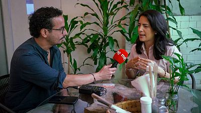Claudia Ortiz, opositora salvadoreña, es entrevistada por un periodista de Radio Nacional de España en un estudio con plantas. Se observa un micrófono "rne rne" y un grabador de audio sobre la mesa.