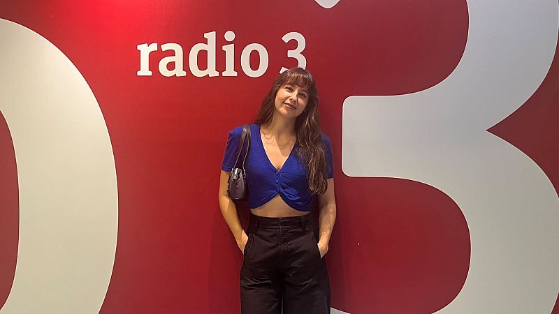 Laura Pardo, presentadora de Radio 3, con top azul eléctrico y pantalones negros, frente a pared roja con logo de Radio 3.