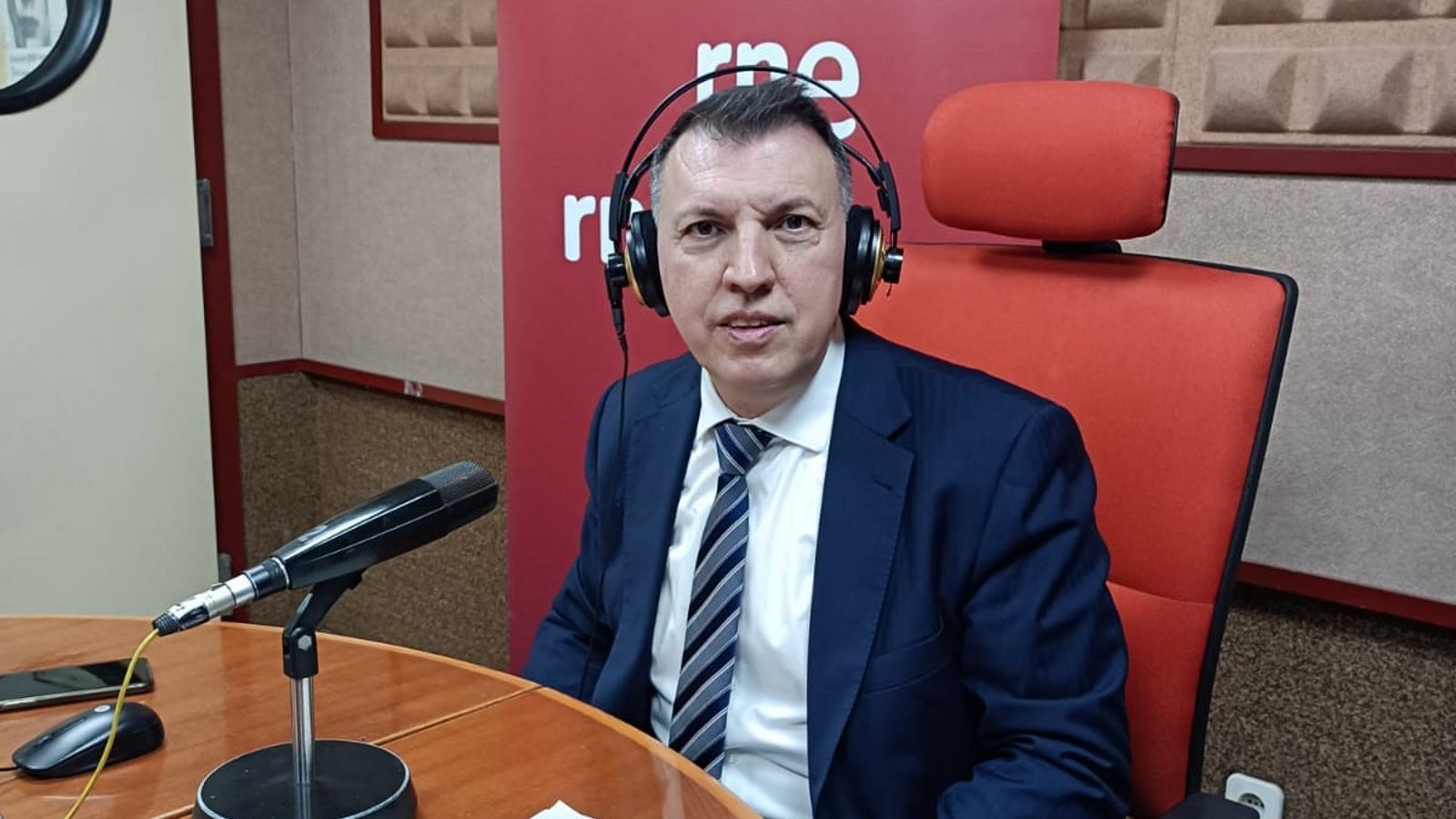 Íñigo Alfonso, presentador, en un estudio de radio con traje azul, camisa blanca y corbata. Auriculares, micrófono, móvil y ratón sobre la mesa. Fondo con logo de RNE.