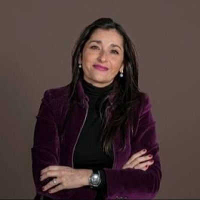 Carla Avilés, con cabello oscuro y liso, viste chaqueta morada y jersey negro. Brazos cruzados, pendientes de perlas y reloj plateado. Fondo liso marrón.