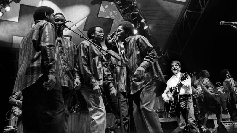 Paul Simon toca la guitarra ac�stica mientras Ladysmith Black Mambazo canta en micr�fonos en un escenario, en una actuaci�n en blanco y negro.