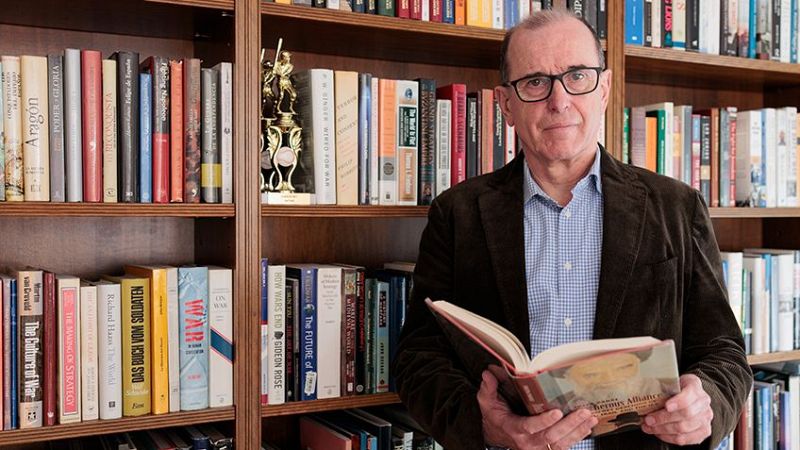 Salvador Sánchez, con gafas y camisa de cuadros, sostiene un libro de alquimia frente a una estantería llena de libros.