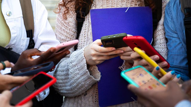 Manos de diversos tonos de piel interactúan con teléfonos móviles de distintas fundas, con pantallas encendidas. Se aprecian detalles de vestimenta y accesorios como un lápiz óptico y un anillo.