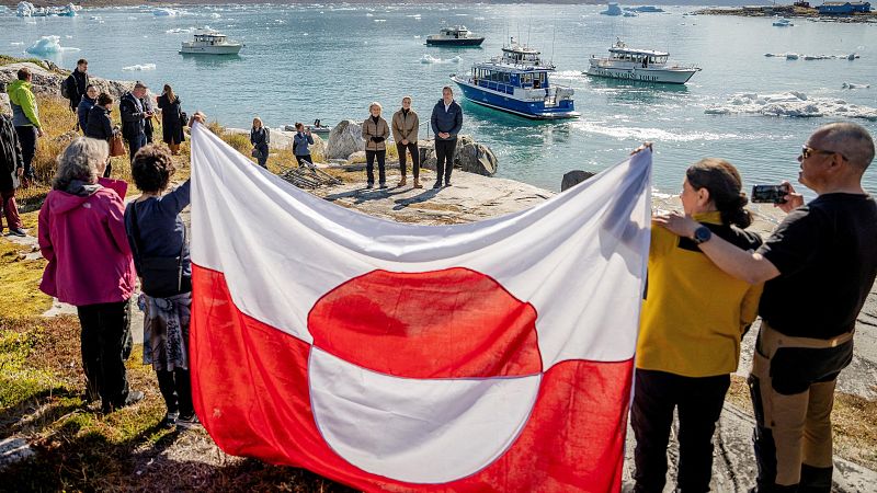 Personas sostienen la bandera de Groenlandia frente a icebergs, con Mette Frederiksen, Kim Kielsen y Mike Pompeo presentes. Embarcaciones navegan en el mar.