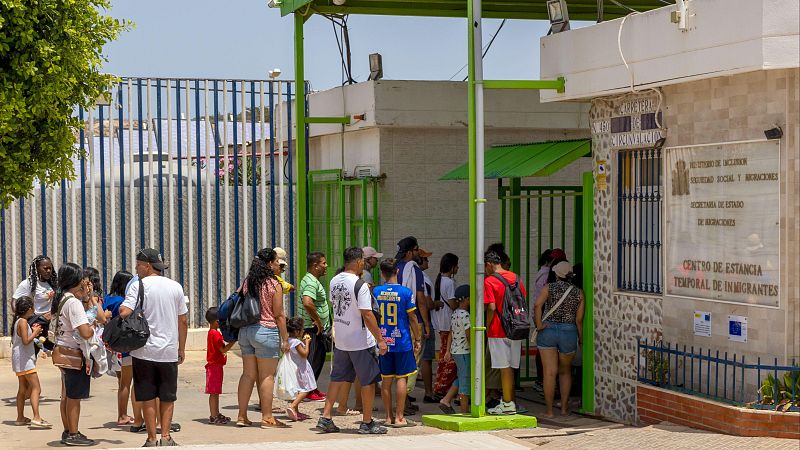 Exterior diurno del CETI de Melilla. Un grupo de 20-30 personas, incluyendo niños, espera frente al edificio con fachada clara y entrada metálica verde. Se aprecian carteles de identificación y logos de la UE.