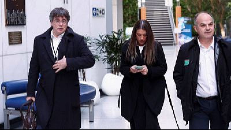 Carles Puigdemont, una mujer y Jordi Turull transitan por un interior moderno. Puigdemont, con gafas y abrigo, lleva un bolso. La mujer observa un objeto, y Turull, con un pin verde, camina a la derecha.