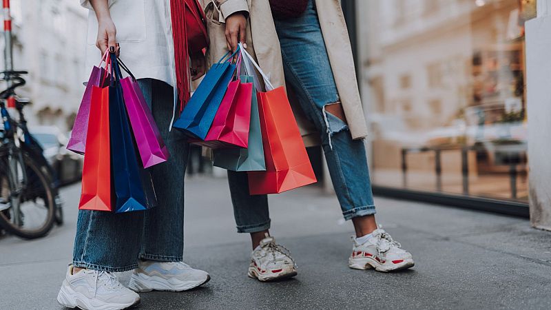 Dos personas con vaqueros y zapatillas, cargadas de bolsas de compra de colores, pasean por una calle urbana con tiendas al fondo.