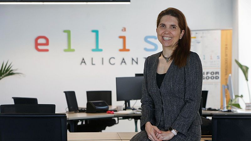 Nuria Oliver, científica informática, sonríe a cámara en una oficina luminosa. Lleva chaqueta de tweed gris oscuro y un collar. Detrás, logo "ellis ALICANTE" y banner "FOR GOOD".