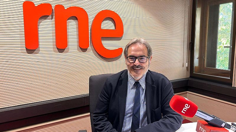 Javier Alenza, director de RNE Navarra, en un estudio de radio con el logo "rne" visible en la pared y en el micrófono. Viste traje y gafas, y sonríe a la cámara.