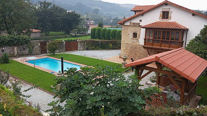 Vista aérea de \"El Caserón del Pas\", propiedad rural con casa tradicional, jardín, piscina y pérgola, rodeada de vegetación y colinas.