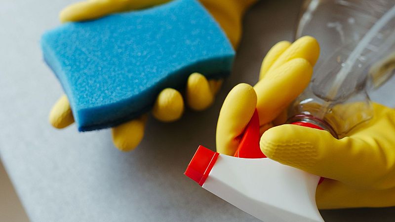 Manos enguantadas de amarillo sostienen esponja azul y pulverizador rojo/blanco sobre superficie clara. Detalle de limpieza.