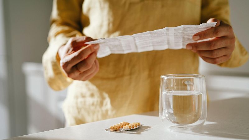 Manos sostienen un folleto de instrucciones junto a un vaso de agua y un blíster de pastillas sobre una superficie clara.