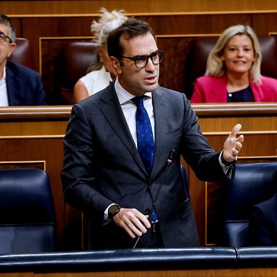 Carlos Cuerpo, Ministro de Economía, habla en el Congreso de los Diputados, con gesto explicativo y gafas, vestido de traje oscuro y corbata azul.
