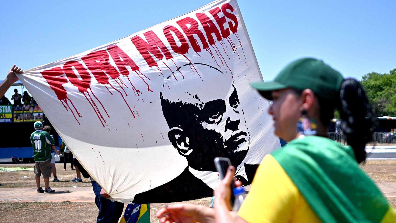 Pancarta de protesta "FORA MORAES" con retrato de Alexandre de Moraes, sostenida por manifestantes con vestimenta que evoca la bandera brasileña.