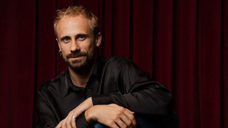 Oriol Pla, actor y director teatral, posa con camisa negra y pendiente plateado ante un telón de terciopelo rojo. Premio Nacional de Teatro 2026.