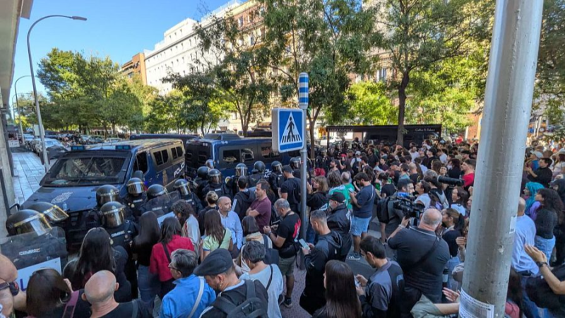 Manifestaci�n en calle urbana con multitud y polic�a antidisturbios en formaci�n de contenci�n. Se aprecian furgonetas policiales al fondo.
