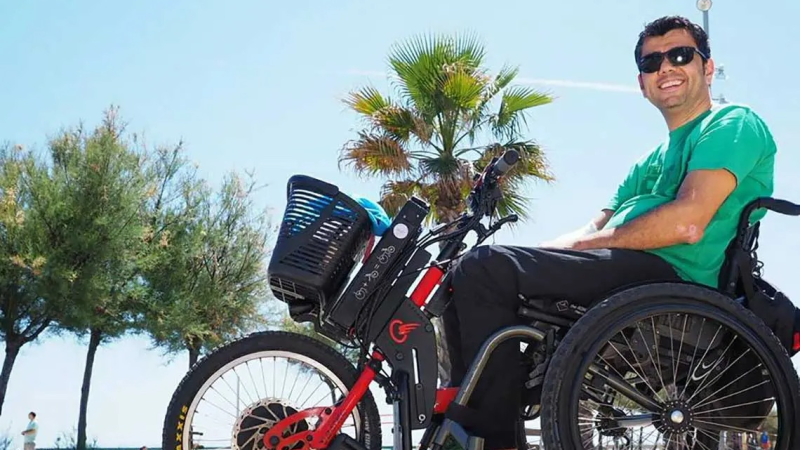 Para-atleta espa�ol Javier de O�a, sonriente en su silla de ruedas con acople el�ctrico, bajo un cielo azul y palmeras.