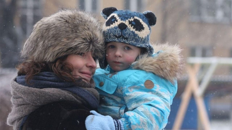 Mujer con gorro de piel y bufanda abraza a un ni�o con gorro de mapache azul en un d�a nevado. El ni�o mira al frente con seriedad.
