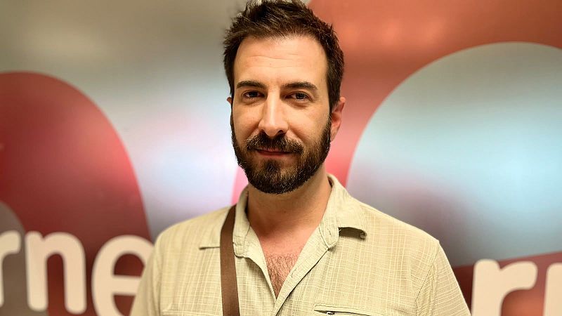 Hombre de mediana edad con barba y bigote oscuros, camisa beige y correa cruzada, sonríe a cámara. Fondo desenfocado con "rne" en blanco.