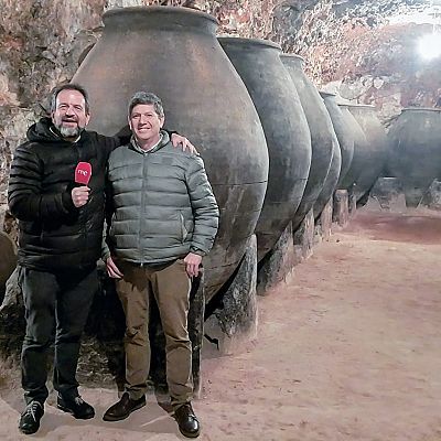 José Ribagorda, con micrófono, y un hombre canoso sonríen en una bodega subterránea con tinajas y barriles de vino. Ambiente rústico y tradicional.