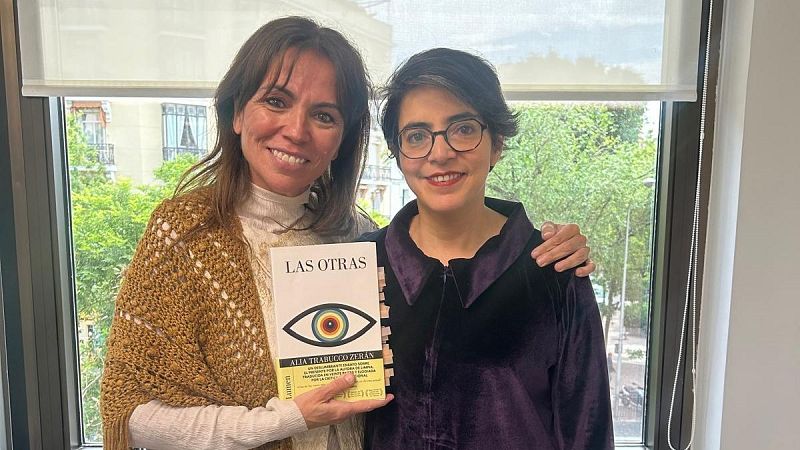 Alia Trabucco Zerán, con gafas y chaqueta morada, posa junto a otra mujer que sostiene el libro "LAS OTRAS" de Lumen. Ambas sonríen a la cámara en un interior con luz natural.