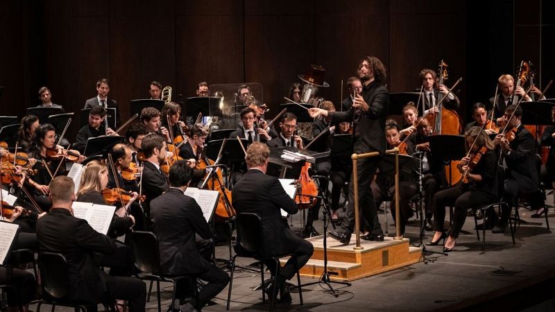 Director joven con cabello rizado dirige orquesta sinf�nica en escenario iluminado. M�sicos de cuerda y viento tocan concentrados. Fondo oscuro.