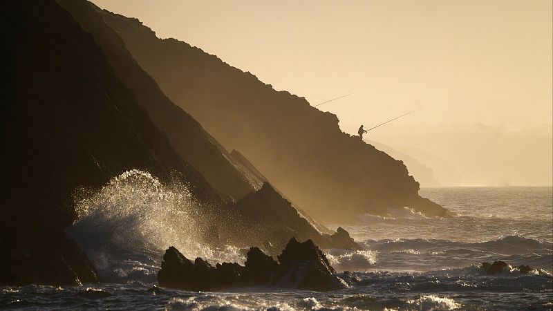 Silueta de pescador con dos cañas en acantilado rocoso, olas rompiendo con espuma dorada por la luz del amanecer/atardecer.