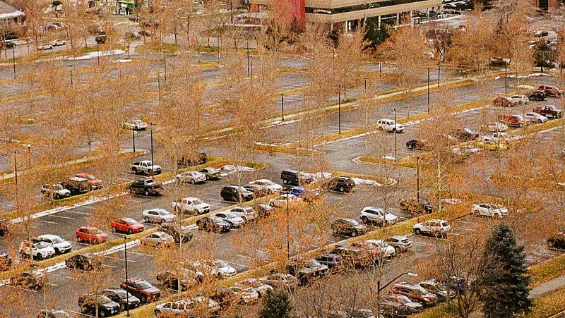 Vista a�rea de un gran aparcamiento con muchos coches, �rboles desnudos y posibles restos de nieve, bajo un cielo nublado o luz tenue, con edificios al fondo.