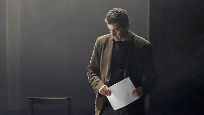 Juan Mayorga, dramaturgo espa�ol, concentrado mirando una hoja de papel en un escenario oscuro y minimalista con iluminaci�n dram�tica.