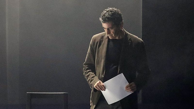 Juan Mayorga, dramaturgo español, concentrado mirando una hoja de papel en un escenario oscuro y minimalista con iluminación dramática.