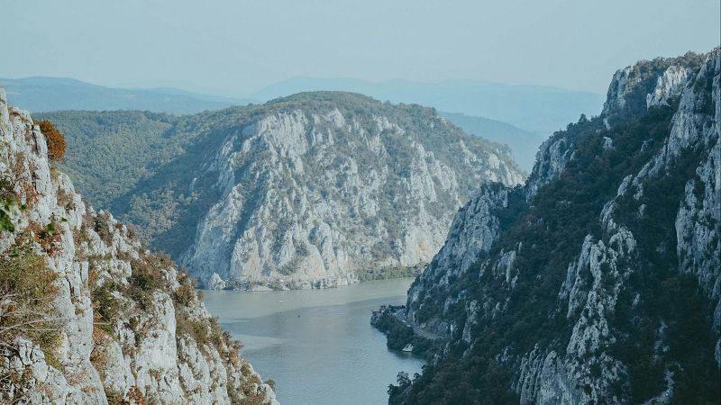 Paisaje de ca��n con acantilados grises, r�o gris verdoso, vegetaci�n oto�al y una carretera en la orilla. Cielo azul p�lido.
