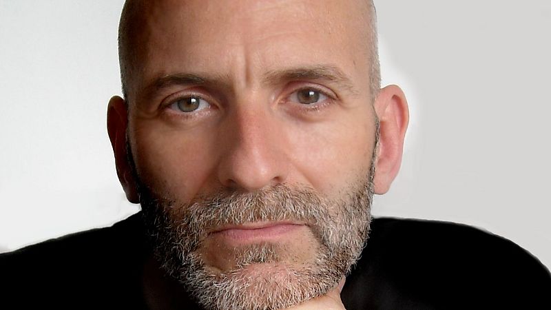 Alejandro Palomas, escritor español, en un retrato de primer plano con barba grisácea y mirada seria. Su mano derecha reposa bajo su barbilla.