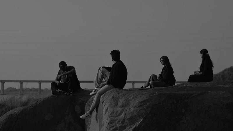 Siluetas de cuatro j�venes sentados en rocas, observando un paisaje con un puente al fondo. Fotograf�a en blanco y negro.