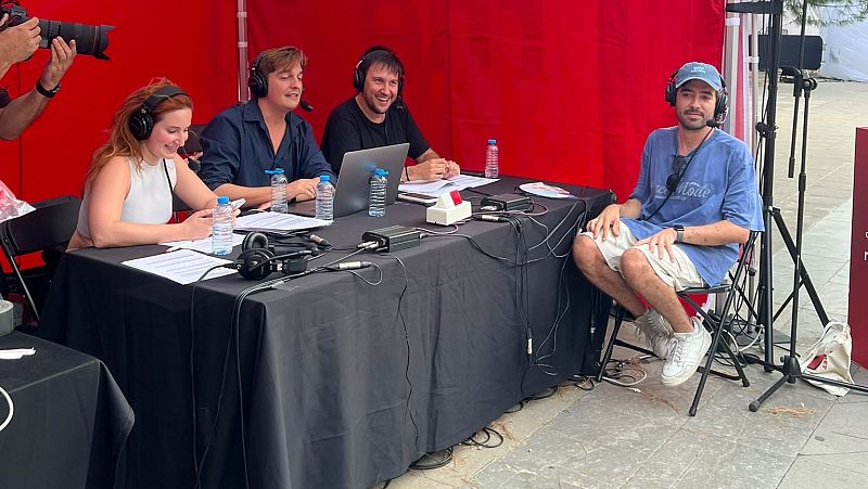 Cuatro personas participan en una grabaci�n de radio o podcast al aire libre, equipados con auriculares y micr�fonos, frente a una mesa con equipo de audio y un tel�n de fondo rojo.