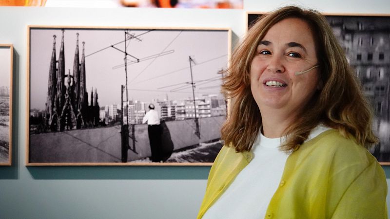 Ada Colau, sonriente y con un cárdigan amarillo, posa frente a fotografías, incluyendo la Sagrada Familia.