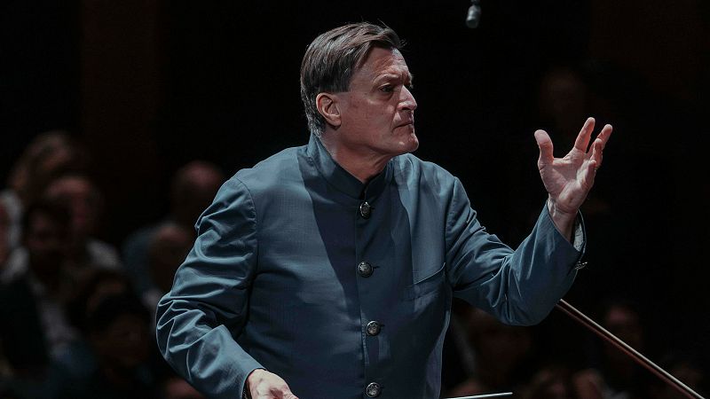 Primer plano de Christian Thielemann dirigiendo con seriedad y pasi�n. Su brazo derecho est� levantado en un gesto de direcci�n orquestal. Fondo oscuro y desenfocado.