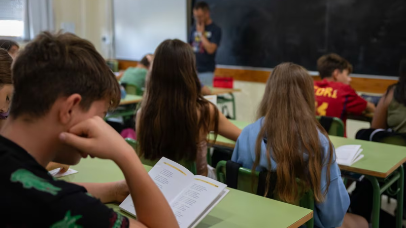Aula escolar con estudiantes leyendo y un profesor de pie. Un joven en primer plano, de perfil, lee un libro abierto con concentraci�n.