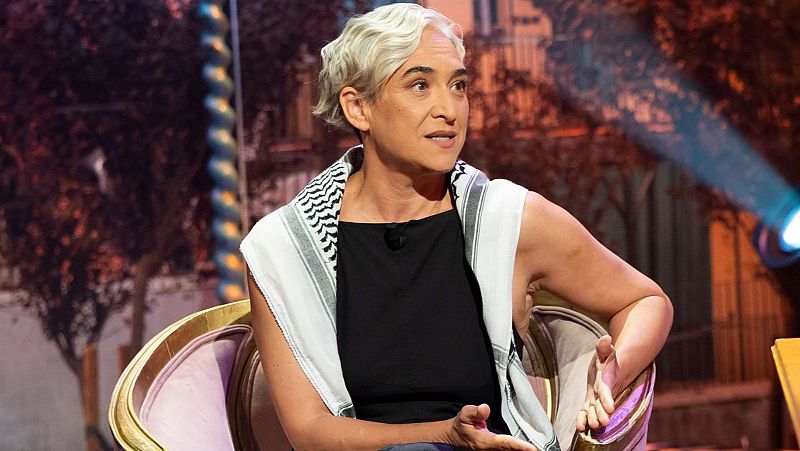Cristina Fallarás, periodista, sentada en sillón de estudio de TV. Viste camiseta negra, pañuelo blanco con patrón geométrico negro. Fondo urbano nocturno desenfocado con luces cálidas y haz azul.