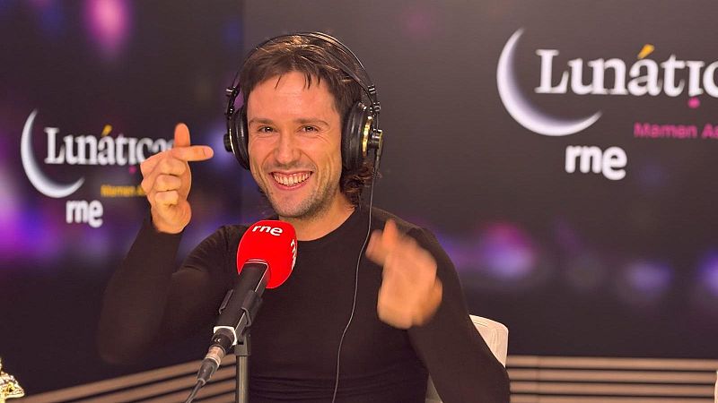 Primer plano de Dani Mateo en estudio de radio, sonriendo con auriculares negros y camiseta negra, haciendo un gesto con las manos. Micrófono rojo "rne" y logos "Lunáticas" y "Mamen As" al fondo.